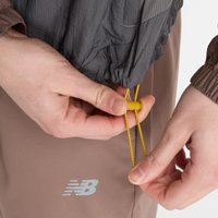 Chaqueta ligera gris con cierre de cordón, sostenida por manos que ajustan un toggle amarillo. Presenta el logo de New Balance en pantalones marrones.
