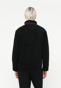 Black fleece jacket with a high collar, textured surface, and a relaxed fit. Features a single back pocket at the neck.