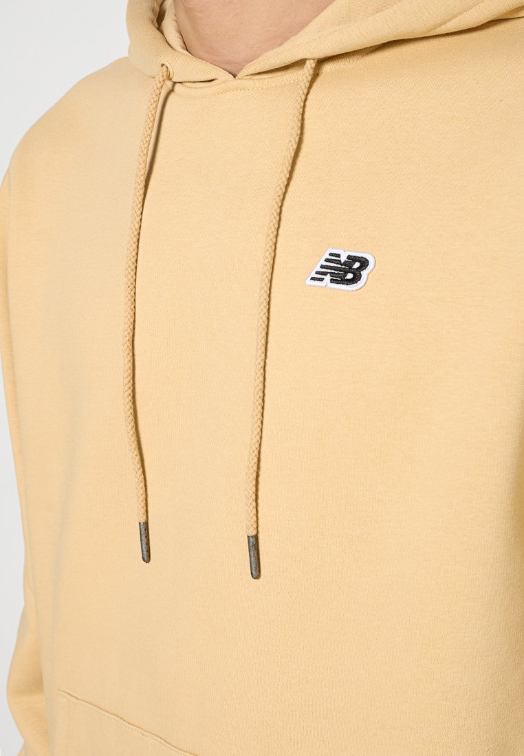 Sudadera amarilla con bolsillo frontal, capucha con cordón y logo negro de New Balance en el pecho. Tejido suave con una textura lisa.