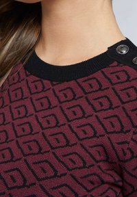 Burgunderfarbener Strickpullover mit geometrischem schwarzem Muster, runden schwarzen Rippkragen und dekorativen schwarzen Knöpfen an der Schulter.