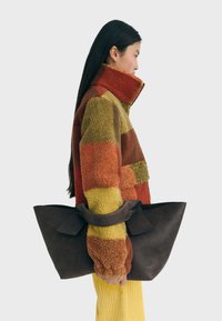 Mujer con cabello largo y oscuro, vistiendo una chaqueta de forro polar de patchwork multicolor y pantalones amarillos, sostenido un gran bolso de mano marrón oscuro a su lado.