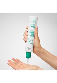 Tube blanc avec accents turquoise, distribuant du shampooing clair dans une paume ouverte. Contient du texte indiquant le type de produit et la marque. Texture lisse.