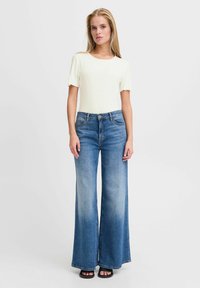 Weißes, geripptes Kurzarmoberteil, kombiniert mit hoch taillierten, weit geschnittenen blauen Jeans. Glatter Stoff mit subtiler Textur und ausgewaschenem Finish. Schwarze Sandalen.
