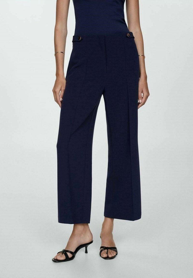 Pantalons larges bleu marine avec une texture lisse, dotés de plis avant et d'accents en métal doré. Associés à des sandales noires à brides.