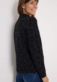 Veste à imprimé léopard noir et gris avec des manches longues, un col rond et une texture douce. Coupe décontractée associée à un jean bleu.