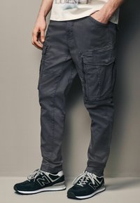 Grå cargo bukser med flere sidelommer, lavet af holdbart stof. Kombineret med mørke sneakers med hvide og sølvfarvede detaljer.