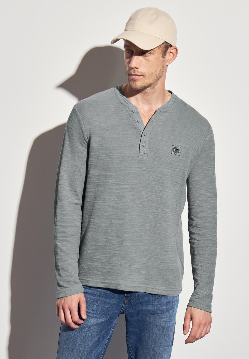 Pitkähihaisen henley-paidan väri on vaalea harmaa, ja siinä on kolmipainikkeinen nappautus, teksturoitu kangas ja pieni neliölogo. Yhdistetty sinisiin farkkuihin.