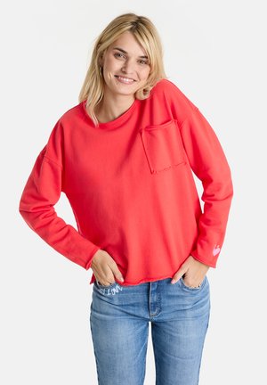 Blonde woman smiling with hands in pockets wearing a bright red long-sleeve shirt with chest pocket and light blue jeans.