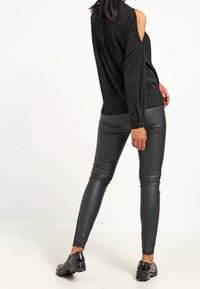 Camicia nera con spalle scoperte e bottoni, abbinata a leggings neri attillati in similpelle e scarpe nere, caratterizzata da una texture liscia e raffinata.