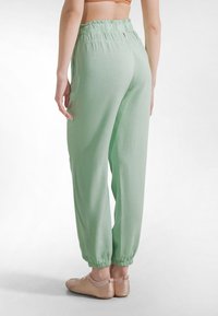 Pantalones jogger de color verde claro, de corte holgado, con cintura elástica y tobillos con puños, combinados con calzado beige tipo slip-on. Tejido suave y texturizado.