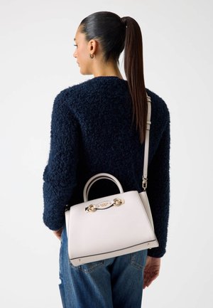 Borsa a mano in pelle bianca con dettagli in oro, design strutturato, manici superiori e tracolla removibile. Indossata con un maglione blu navy.