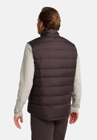 Bruine puffer vest met een gewatteerd ontwerp, hoge kraag en mouwloze stijl, gedragen over een lichtgrijze lange mouwen shirt. Gladde textuur.