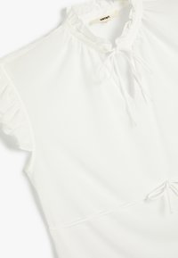 Blusa bianca con colletto arricciato, chiusura a fiocco e maniche corte arricciate. Realizzata in un tessuto a costine con un design aderente.
