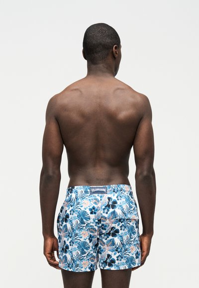Άνδρας μοντέλο φαίνεται από πίσω, φοράει μαγιό σορτς με μπλε και λευκό φλοράλ σχέδιο και ετικέτα Vilebrequin στη μέση.