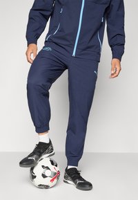 Marineblå træningsdragt med lyseblå accenter; slim-fit joggers med elastiske manchetter, parret med sorte fodboldstøvler og en mønstret fodbold.
