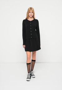 Cárdigan negro de manga larga con escote redondo y detalles de botones, combinado con una falda negra plisada y calcetas negras hasta la rodilla con zapatillas deportivas.