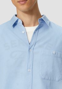Hellblaues Hemd aus strukturiertem Stoff. Verfügt über eine Brusttasche, einen weichen Kragen und zwei Knöpfe auf der Vorderseite.