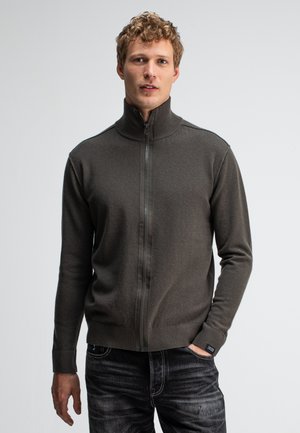 Pull zippé en gris foncé, avec un col haut et des poignets côtelés. Texture douce avec une coupe ajustée. Porté par-dessus un jean foncé.