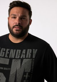 Černé tričko s kulatým výstřihem a velkým, texturovaným šedým nápisem, který nápadně zobrazuje slova "LEGENDÁRNÍ" a "DENIMOVÉ OBLEČENÍ".