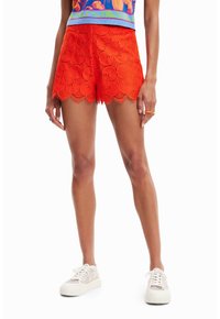Shorts en dentelle orange vif, ornés de découpes florales et d'un ourlet festonné. Associés à des baskets compensées de couleur claire.