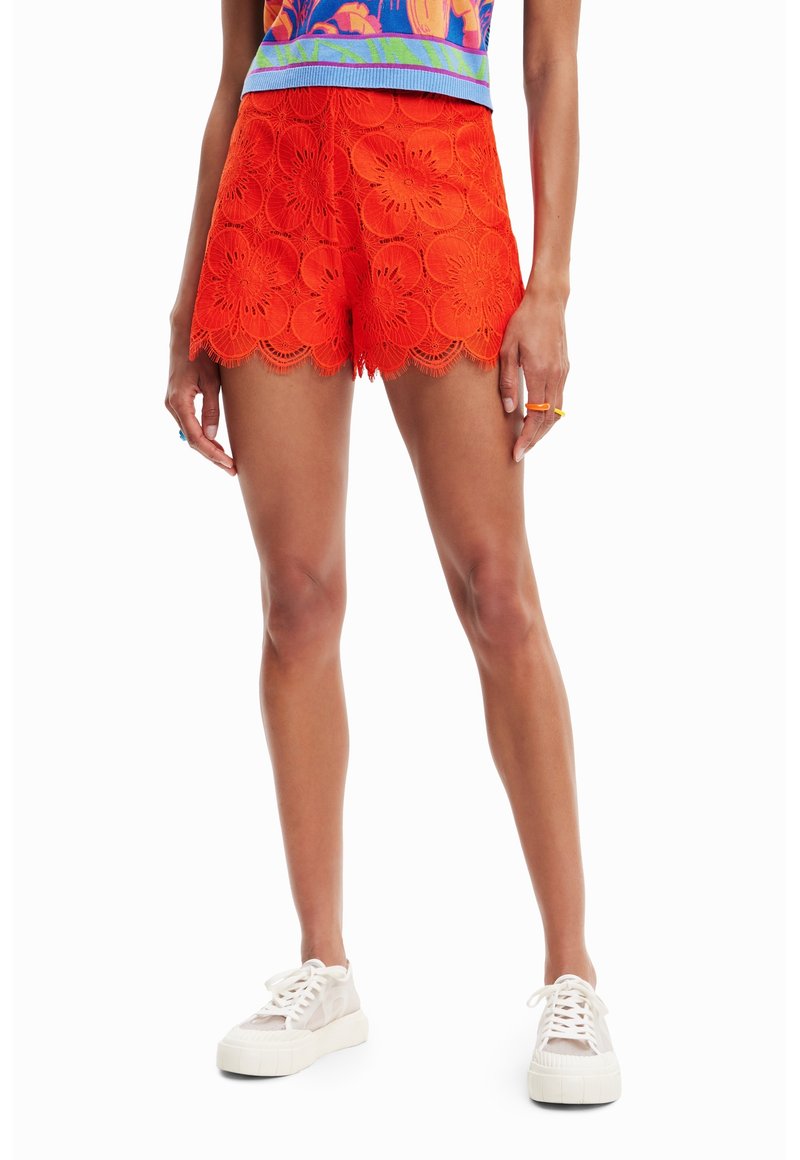 Shorts en dentelle orange vif, ornés de découpes florales et d'un ourlet festonné. Associés à des baskets compensées de couleur claire.