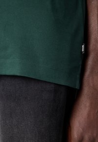 Zelené bavlnené tričko s hladkým povrchom, so zošitým lemom a malou čiernou nálepkou "BOSS" na boku, doplnené tmavými džínsami.