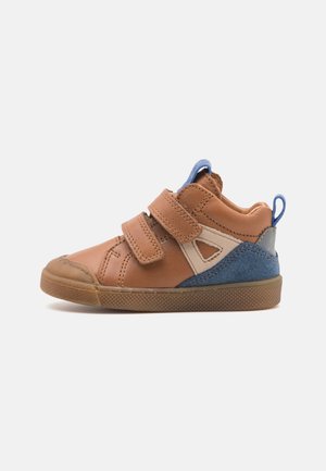 Zapatilla deportiva para niño pequeño en tonos marrón y azul con dos tiras de velcro, detalles laterales beige y suela de goma marrón sobre fondo blanco.