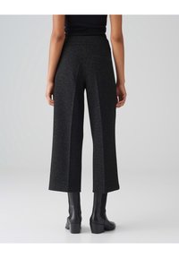 Pantalons larges noirs en tissu texturé, comportant une taille haute, des poches arrière et associés à des bottines noires brillantes.