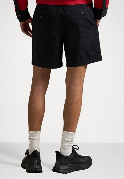 Schwarze Baumwollshorts mit geradem Schnitt und Taschen auf der Rückseite. Getragen mit schwarzen Sportschuhen und weißen gerippten Socken.