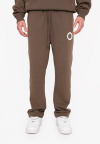 Brune joggerbukser lavet af blødt stof, med en elastisk talje og snor, samt et rundt logo-patch på venstre lår.