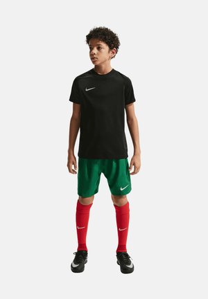 Ragazzo in piedi con maglietta sportiva nera, pantaloncini verdi, calzini rossi al ginocchio e scarpe sportive nere su sfondo uniforme.