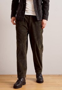 Chaqueta de pana negra, camiseta blanca y pantalones de pierna ancha en verde oliva. Botas negras con cordones. Tejido texturizado con patrones sutiles.