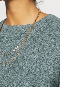 Vaaleanvihreä neulepaita, jossa on avara kaula-aukko ja teksturoitu pinta. Kerrokselliset kultaketjut roikkuvat kaula-aukossa.