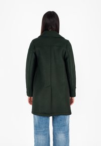 Cappotto verde in lana con colletto a punte, maniche lunghe, due spacco posteriori e taglio dritto, abbinato a jeans a gamba larga blu chiaro.