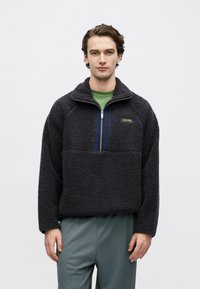 Μαύρο πουλόβερ από φλις με γιακά μισό φερμουάρ, υφή στην επιφάνεια, μπλε πάνελ με χρώμα ναυτικό και αντίθετο κολαριστό λογότυπο· φορεμένο πάνω από πράσινο πουκάμισο.