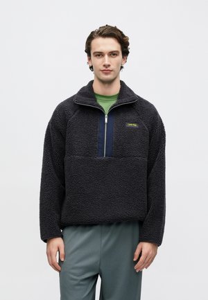 Mustast fliisist pullover poollukuga kraega, tekstuurse pinnaga, mereväe sinise aktsendipaneeli ja kontrastse logomärgiga; kantud rohelise särgi peal.