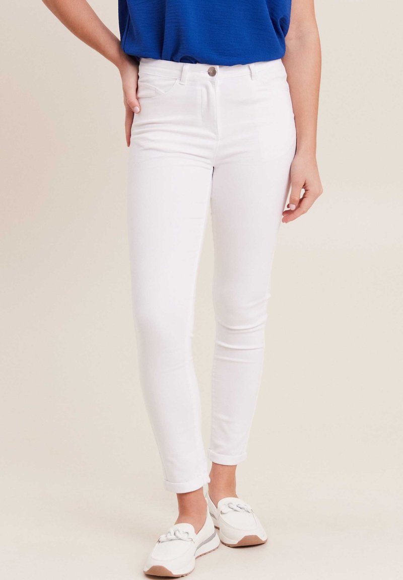 Witte skinny jeans met een gladde textuur, vijf zakken, en opgerolde mouwen, gecombineerd met witte instappers en een blauwe top.