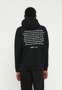 Melns džemperis ar kapuci, priekšpusē kabata un regulējama kapuce. Balts uzraksts aizmugurē. Ribām apdarinātas aproces un apakša. Mīksta tekstūra.