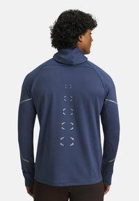 Maglietta sportiva a maniche lunghe blu navy con colletto alto, tessuto lavorato, dettagli riflettenti e decorazioni circolari lungo la schiena.
