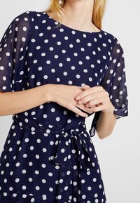 Navyblå klänning med vita prickar, korta genomskinliga ärmar, rund halsringning och knytdetalj i midjan. Tyget verkar lätt och slätt.