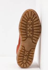 Semelle d'une chaussure marron à semelle en caoutchouc avec des rainures profondes, positionnée à la verticale contre une surface blanche.