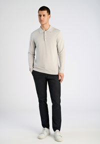 Helle graue Langarmpoloshirt mit Kragen und einer Drei-Knopf-Leiste, kombiniert mit dunklen Hosen und weißen Sneakers, vor einem schlichten Hintergrund stehend.