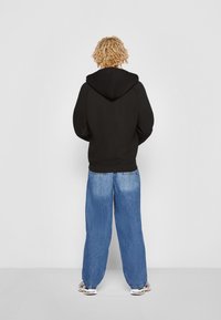 Sudadera negra con capucha ajustable y puños acanalados, combinada con vaqueros de denim sueltos de color azul claro. Vista desde la parte trasera.