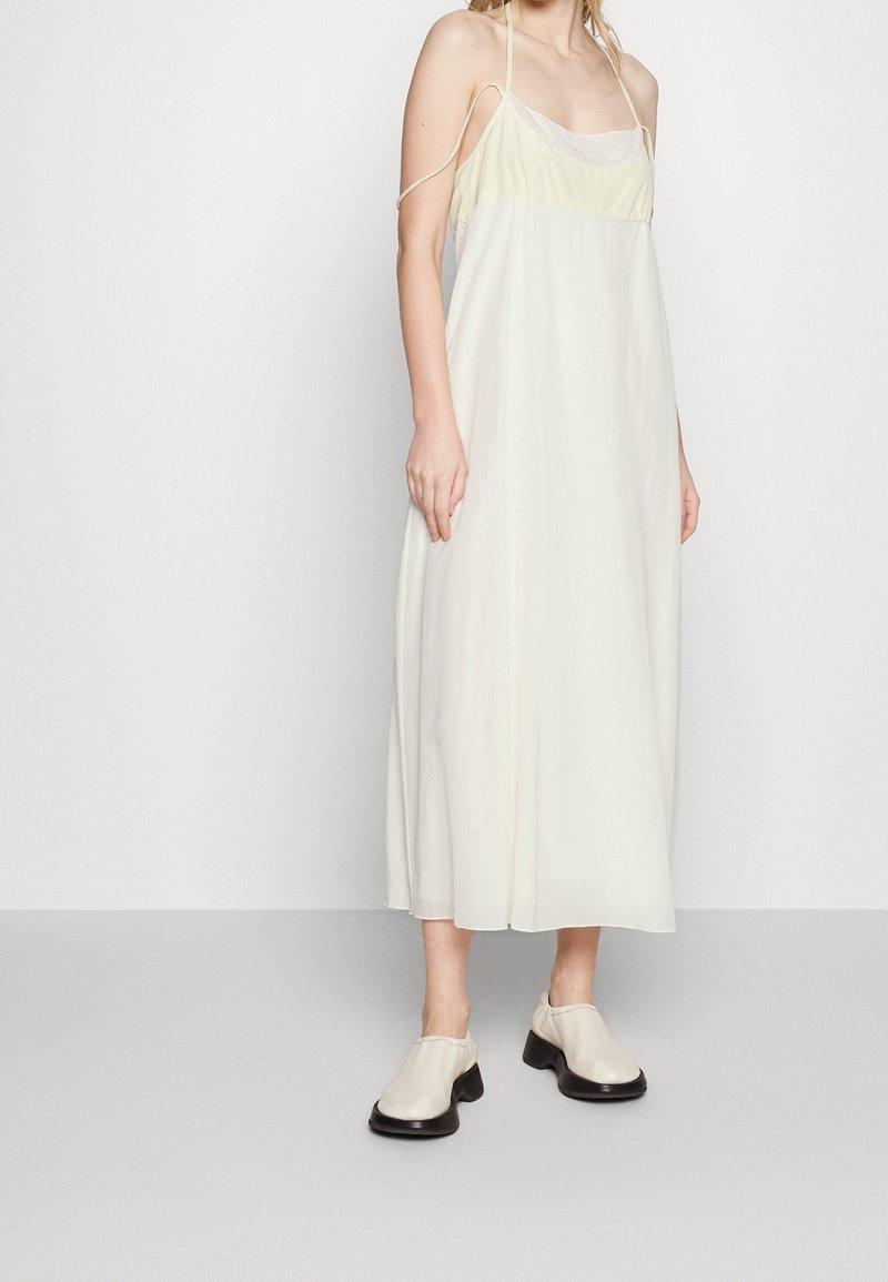 Robe maxi sans manches de couleur jaune clair, avec une coupe décontractée et des fines bretelles ; associée à des chaussures à semelles épaisses et aux tons neutres.