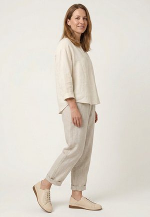 Frau trägt beige Leinen-Langarmshirt, passende Leinenhose, am Knöchel hochgekrempelt, und bequeme beigen Schnürschuhe, steht seitlich und lächelt.
