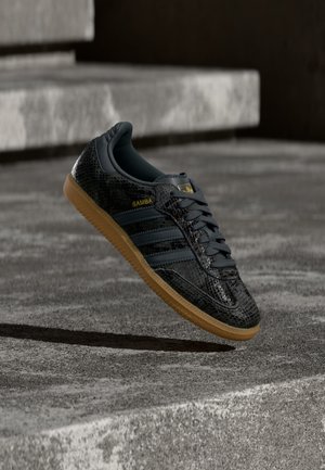 Svart Adidas Samba-sneaker med teksturert mønster, såle i gummi og gullfarget logo, plassert i luften over betongtrinn.