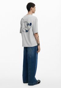 Szürke, oversized póló, amelyen egy nagy hímzett Mickey Mouse sziluett látható a hátsó részen. Kék, széles szárú farmerral párosítva.
