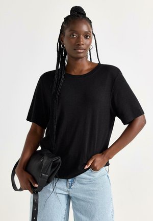 Kvinde med lange fletninger iført en sort t-shirt og lyseblå jeans, der holder en sort håndtaske og står foran en ensfarvet baggrund.