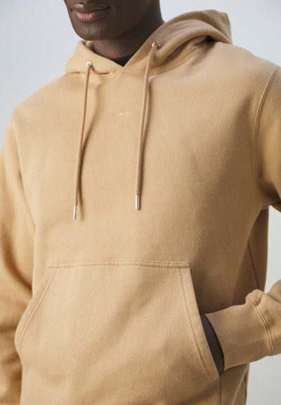 Hanorac cu glugă de culoare bej, dintr-un material moale. Are un buzunar frontal, șnur cu capete din argint și un logo minimalist pe piept.