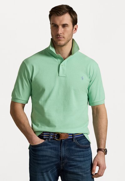 Polohemd aus mintgrüner Baumwolle mit einem Kragen mit zwei Knöpfen, kurzen Ärmeln und einem gestickten Logo auf der linken Brust. Getragen mit blauen Jeans und einem mehrfarbigen Streifen-Gürtel.